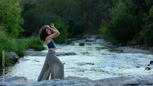 A Serene Moment by the River: A Woman with Curly Hair and Flowing Pants Posing Elegantly Against the Backdrop of Tranquil Water and Lush Greenery