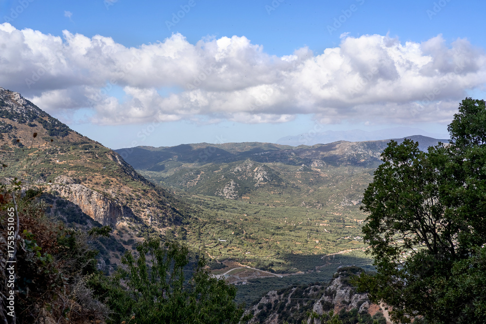 Naklejka premium Beautiful panoramic view of the Cretan countryside with mountains, green valleys, olive groves, and a small traditional village under a bright blue sky with scattered clouds. Captured on a sunny day