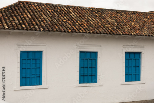 Papier peint A serene colonial-style wall with vibrant blue windows stands beneath a weathered clay tile roof, evoking cultural heritage, simplicity, and timeless architectural charm