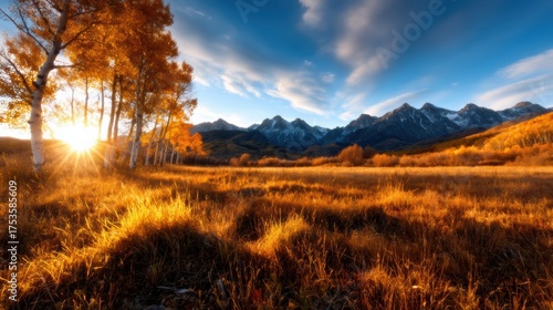 This stunning landscape captures a golden sunset casting warm light over a field of grass with vibrant autumn trees and dramatic mountain peaks in the background.