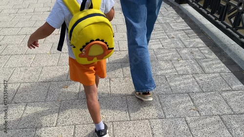A young child with a bright yellow dinosaur backpack and orange shorts walks alongside an adult on a sunny, paved path.