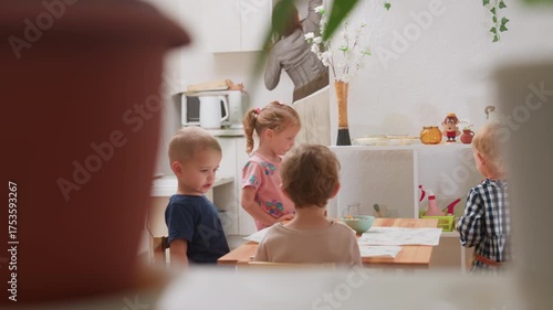 Children engaged around table while instructor reaches shelf and hands book to girl in pink clothing during kindergarten activity with classmates observing showing cooperation learning responsibility