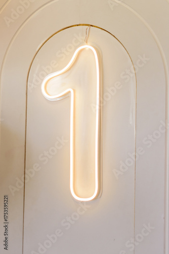 A stylish modern neon sign of the number one, glowing with warm light on an arched beige wall, perfect for a first birthday party decoration.