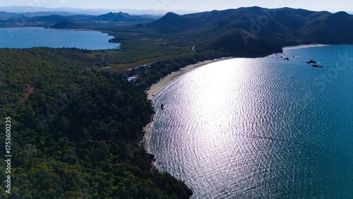 Fotografia Breathtaking aerial view of a pristine, sandy beach surrounded by lush, verdant forest and rugged, towering mountains under a clear, azure sky