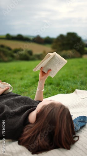 A woman is laying on a blanket reading a book. The scene is peaceful and relaxing, with the woman enjoying her time outdoors