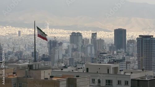 Wallpaper Mural The Iranian flag waves on a tall pole above Tehran skyline, with high-rise buildings, cranes, and mountains in the distance, symbolizing national identity, resilience, and urban transformation. Torontodigital.ca