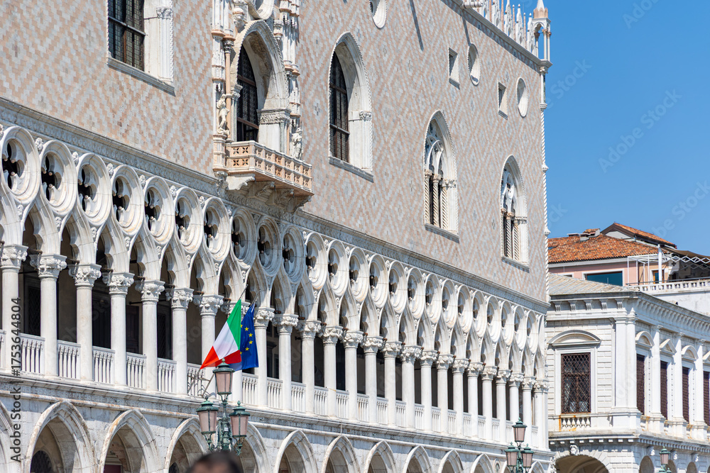 Fototapeta premium Doges Palace Facade with Italian and EU Flags. Close-up of the ornate Gothic facade of Doges Palace in Venice, featuring arched windows, marble columns, and national flags.