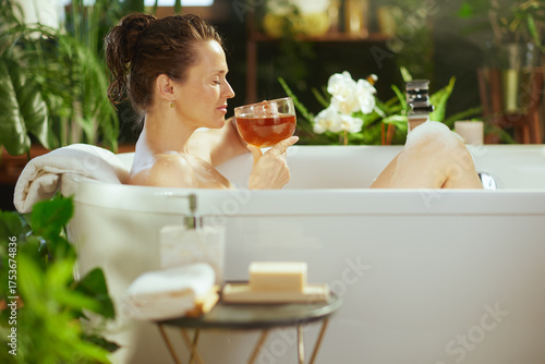 An adult woman, eyes closed, enjoys tranquility in a white bathtub, holding tea. Lush green plants & soft lighting enhance this serene, spa-like self-care experience.