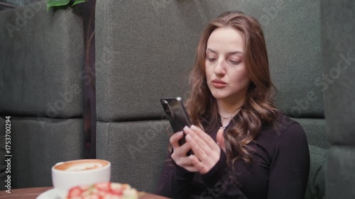 Young woman in black outfit seated in green booth fixated on smartphone with dessert and cup of latte on wooden table. Surrounded by lush indoor plants, and books