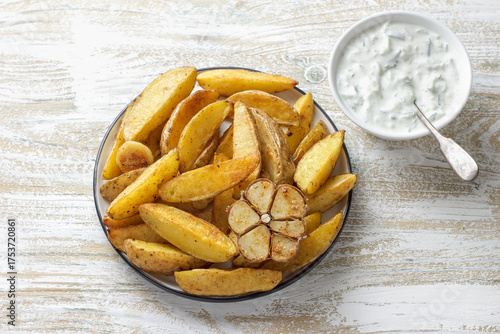 Golden baked potato wedges with garlic, spices and Greek tzatziki sauceon rustic plate. Tasty homemade snack or side dish, healthy vegetarian food