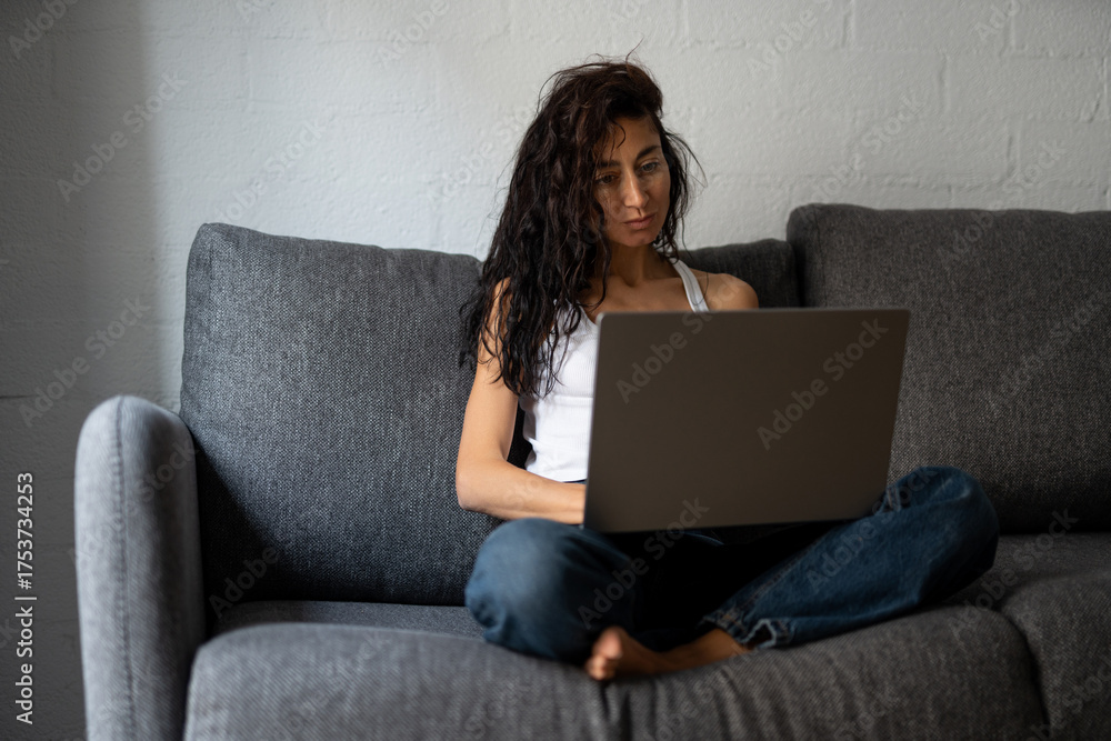 Fototapeta premium Freelance woman working from home on laptop, sitting comfortably cross-legged on a couch near window, wearing casual clothes and concentrating on her remote tasks.