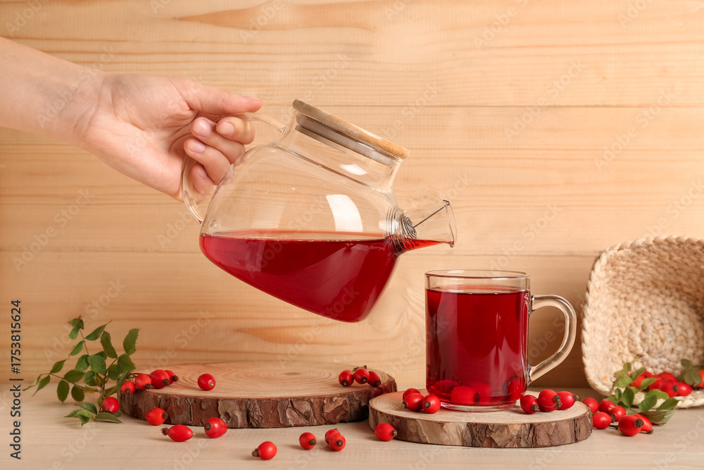 Fototapeta premium Woman pouring hot rose hip tea from teapot into glass cup on wooden background