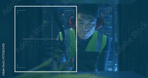 Kneeling Korean technician inspecting tablet in data center, with LEDs server racks safety vest