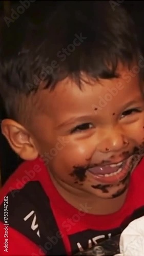 A toddler boy, covered in chocolate cake around his mouth and cheeks, is laughing heartily while wearing a red shirt, enjoying his birthday or a treat.