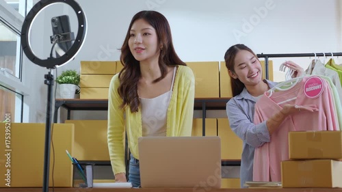 Two young asian woman live streaming online clothing store while showing clothing with smartphone on social at home, entrepreneur streaming for selling online clothing and presenting, online seller.
