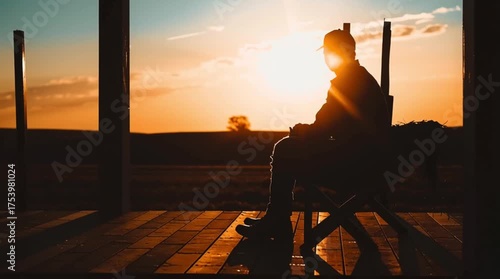 Silhouette of a person sitting on a wooden porch, contemplating a vibrant golden sunset over an open rural landscape