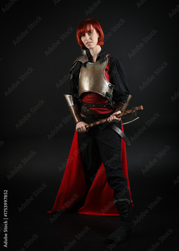 Naklejka premium fantasy portrait of warrior woman knight, model wearing historical medieval armour costume, red fairytale cloak. holding battle axe weapon, isolated dark studio background.