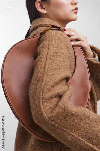 Asian woman wearing a warm brown coat carries a sculpted leather bag over her shoulder, illustrating chic urban fashion and confident style in a minimalist portrait.