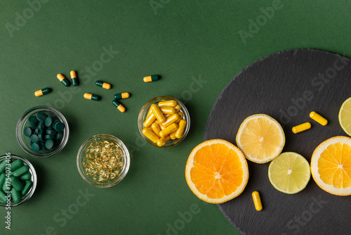 Fototapeta Naklejka Na Ścianę i Meble -  Nutritional supplements, various vitamins and minerals, pills and tablets from above on a black stone desk on green background. Citrus fruits - lemon, orange and lime as a source of natural vitamins.