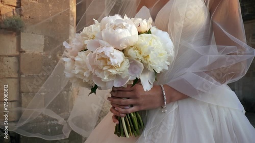 Bride in white gown holding bouquet of white peonies beside stone wall with veil flowing