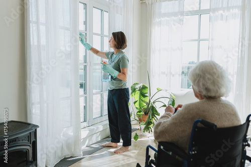 Cleaning service for seniors concept. Female cleaner in gloves washing big panoramic windows in cozy living-room using detergent and talking to old disabled lady home owner in wheelchair