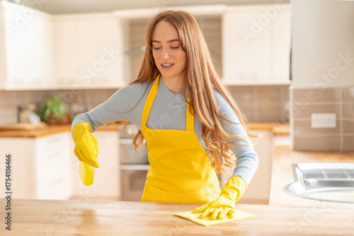 Tableau sur toile Young woman cleaning kitchen countertop while wearing yellow apron and gloves du