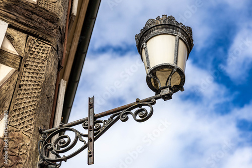 Historische Straßenlaterne an Fachwerkhaus im Elsass – Detailaufnahme von Schmiedeeisen und Holzarchitektur vor blauem Himmel