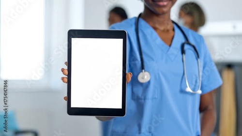 Doctor in blue scrubs, stethoscope, holding up a blank tablet. Medical colleagues blurred