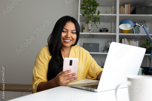 Smiling latin american woman using laptop and smartphone at home office