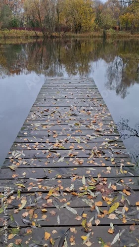 A wooden pier covered with wet autumn leaves leads to a calm lake on an overcast day. A peaceful autumn landscape with trees reflected in the calm water.