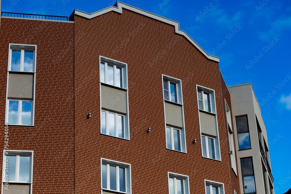 Fototapeta premium A brick house with large windows, white frames, and decorative roof elements. Contemporary style, smooth walls, a bright sunny day, and blue skies.