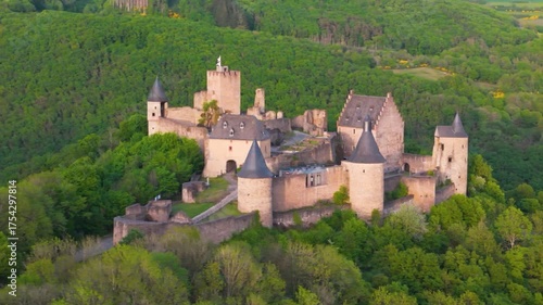 vast area bourscheid castle château de green valley hills behind walls black domed towers flag top foggy day tall trees colorful to left around right drone panning 