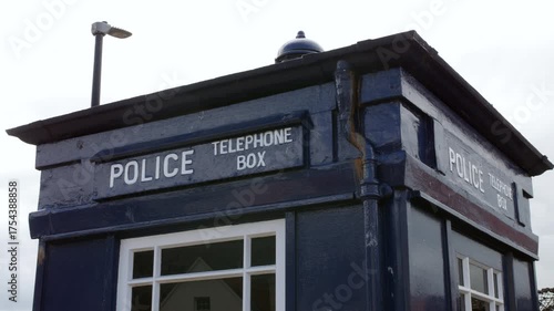 A blue police telephone box standing beneath a cloudy sky, with a mounted security camera and white lettering above the small front window