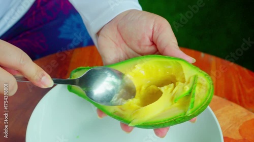 Woman hand scooping avocado flesh using stainless steel spoon to a white plate, in a cozy home environment