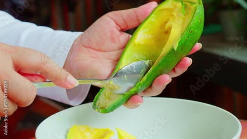 Woman hand scooping avocado flesh using stainless steel spoon to a white plate, in a cozy home environment