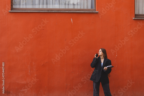 Serious long hair brunette business woman wear jacket, go to job, hold tablet, notebook and have conversation on mobile phone. Busy and successful. Orange wall.