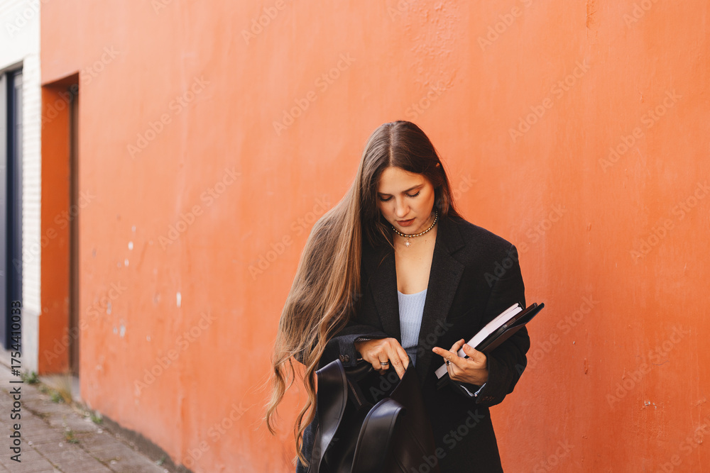 Naklejka premium Elegant young long hair woman looking in her black leather bag her mobile phone or purse. Business style woman wear black blazer and bag on the street. Street style, fashion outfit. Orange wall.