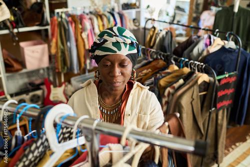 Young adult Black woman browsing clothing rack in thrift shop, selecting garments among assorted secondhand clothes, focused expression, surrounded by vintage apparel displays