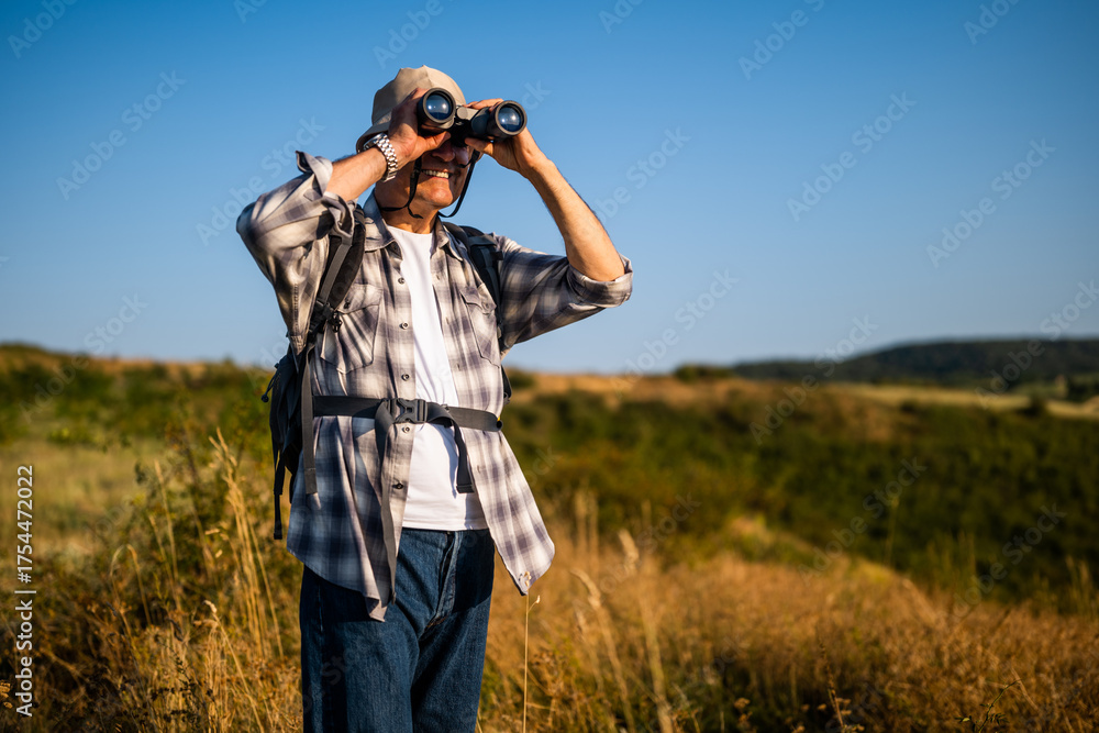 Fototapeta premium Senior man with backpack using binoculars while hiking in nature. 