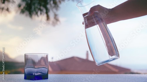Woman Pouring Fresh Water from Jug into Glass Outdoors with Clear Sky and Cozy Home Atmosphere