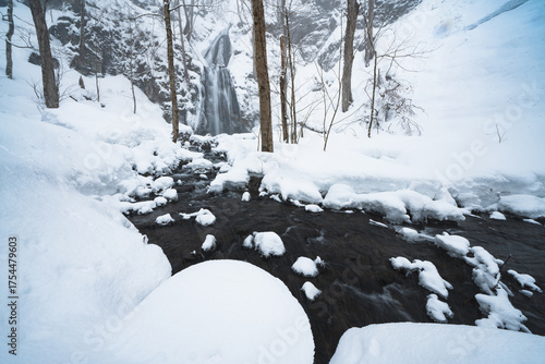Snow-covered waterfall and stream in a quiet winter forest in northern Japan, capturing the beauty of nature surrounded by white snow and cold flowing water.