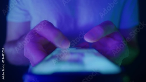 Close-up of hands holding a smartphone and quickly scrolling through social media feeds at night in neon light. Doom scrolling, phone overuse and digital addiction concept. 