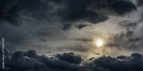 dark dramatic cloudy sky with thick clouds and dim sun backlighting from behind atmospheric haze. Widescreen panoramic photo of natural cloudscape for moody design or creative sky background