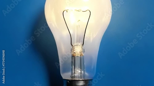 Close-up of a lit bulb, illuminating against a textured blue background. Filament glows brightly. Glass encasement is clear