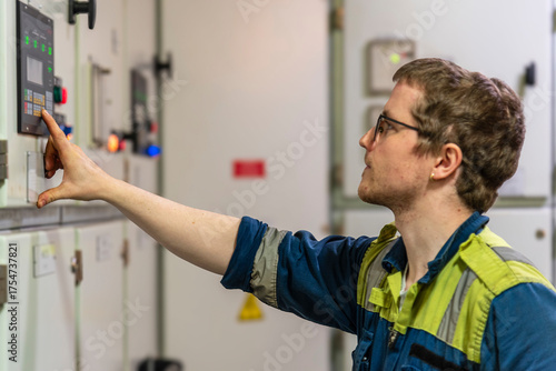 Young male engineer in a blue and yellow safety uniform operates a control panel in a ship's engine room.