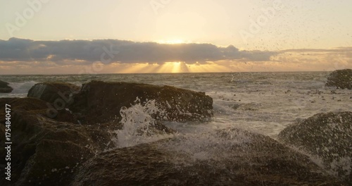 Crashing ocean waves spraying sea spray on jagged shore rocks, with sun rising behind low clouds © vectorfusionart