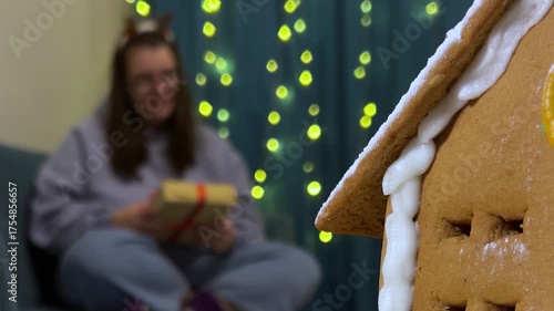 Horizontal defocused video. Woman out of focus sits on sofa in room and gives New Year gifts to husband, spouse, lover, friend. Gingerbread brown house with winter decor in foreground. Bokeh light
