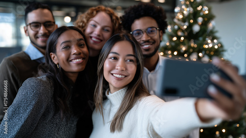 Diverse group of happy young business people or friends taking a selfie photo on smartphone during a christmas party in a modern office.