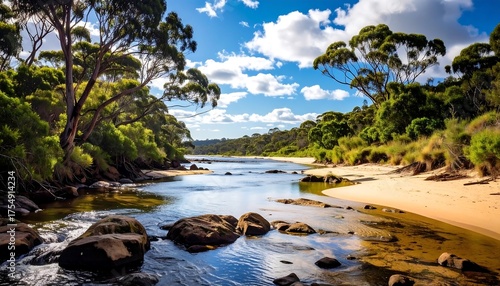 Fototapeta Naklejka Na Ścianę i Meble -  A tranquil river flows toward the sea, fringed by lush green trees. Sunlight dapples the water, reflecting the blue sky with fluffy clouds