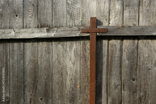 A rusty cross stands against a weathered wooden fence. The wood is aged and textured, creating a rustic backdrop for the cross.